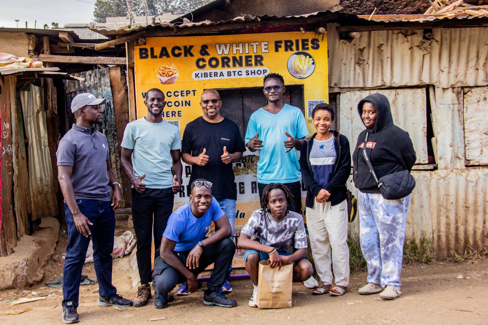 A group outside Black & White Fries Corner, a Kibera shop with a painted sign reading 'We accept Bitcoin here'