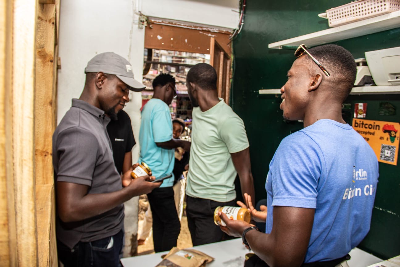 Two young men holding up products under a 'Bitcoin accepted here' Afribit sign in a Kibera kiosk